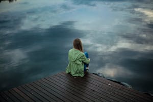 Woman alone Sitting on Wooden Planks in front a lake mirroing the sky