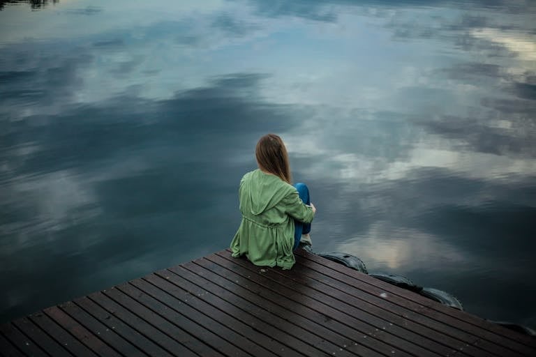 Woman alone Sitting on Wooden Planks in front a lake mirroing the sky